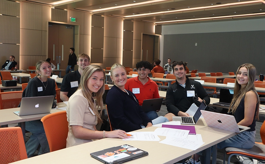 A group of seven students smiling while seated at a conference table with laptops and notebooks in a modern classroom setting.