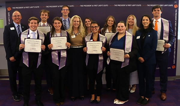 Group of students wearing purple stoles and holding Clemson University certificates, posing with faculty at an awards ceremony backdrop.