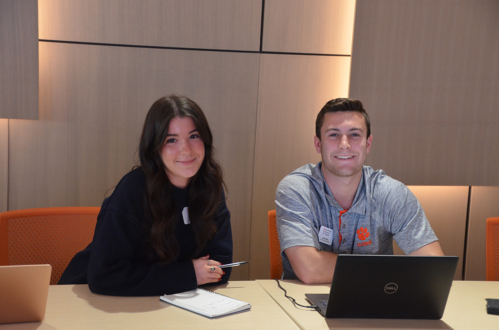 Two students smiling at a conference table, with notebooks and laptops in the background.