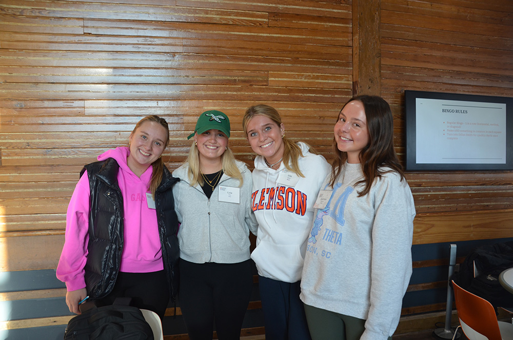 A group of four young women posing together in a casual setting, smiling and wearing athletic clothing and accessories.