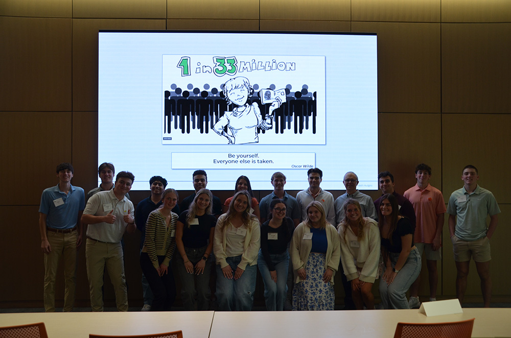 A group of young adults poses together in front of a large screen displaying a cartoon illustration with the text "1 in 33 Million" and the quote "Be yourself. Everyone else is taken."