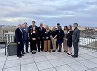 Group of people in formal attire posing together outdoors on a paved terrace under a cloudy sky.