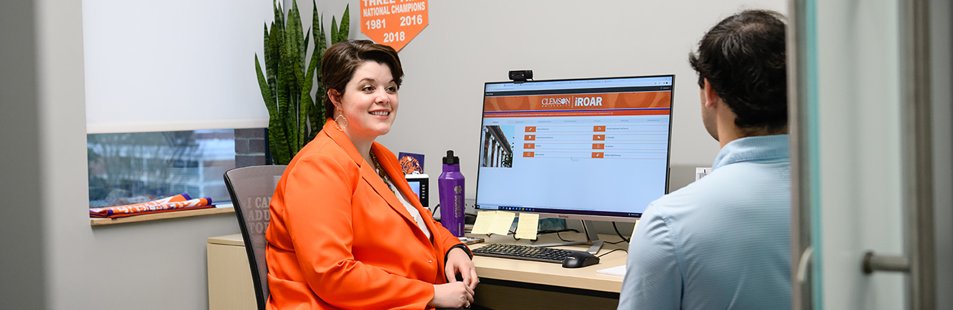 Woman with short dark hair wearing orange jacket talking to male with short brown hair and wearing light blue shirt.