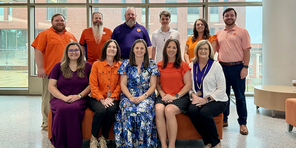 Group photo of faculty and staff in a campus building, posing together in front of large windows.