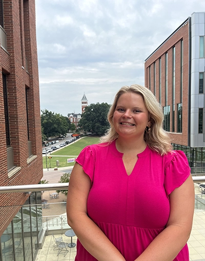 A smiling woman in a pink dress stands on a balcony, with a view of a university campus and classic architecture in the background.