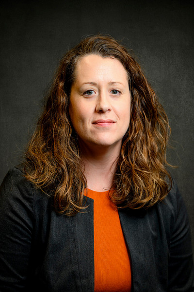 Professional headshot of a woman with curly hair, wearing a black blazer over an orange top.