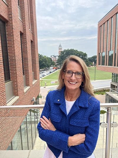 A smiling woman in a blue jacket stands with her arms crossed, overlooking a cityscape with a clock tower in the background.