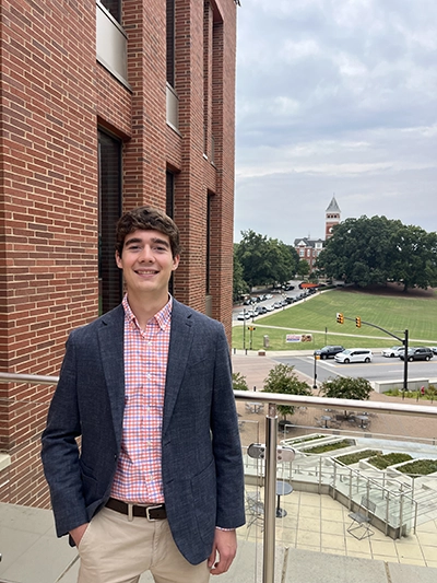 A young man in a blazer and button-up shirt stands on a balcony, smiling at the camera with a university campus in the background.