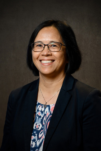 Professional headshot of a woman with short black hair and glasses, smiling in a black blazer over a patterned top.