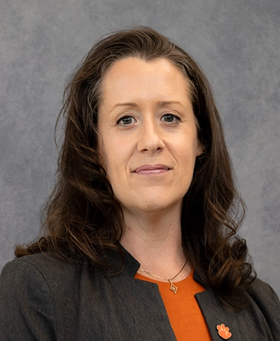 Head-and-shoulders studio portrait of a woman with long brown hair wearing a dark blazer over an orange top, against a gray background.