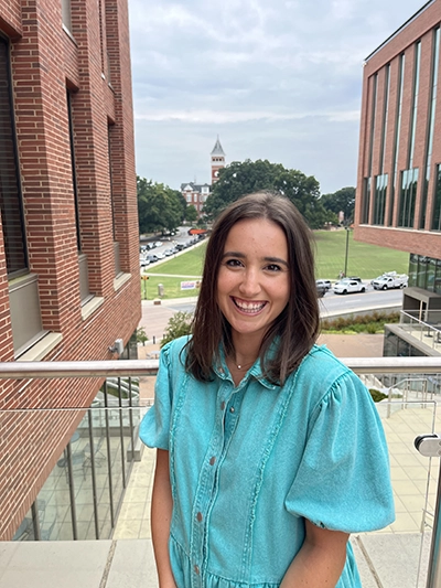 A young woman in a turquoise dress smiles in the foreground, with a university campus and clock tower visible in the background.