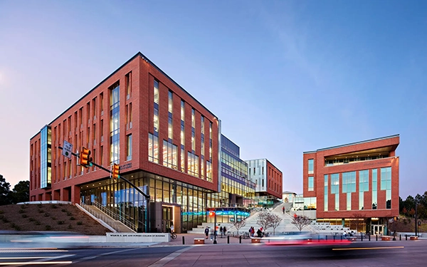 Modern university building complex with large glass windows and a contemporary design, set against a twilight sky.