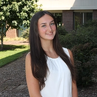 A young woman smiling outdoors, wearing a white blouse, with greenery and a building in the background.