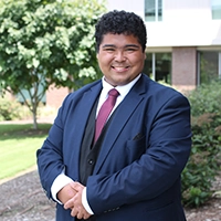A professional portrait of a young man in a suit, smiling confidently outdoors.