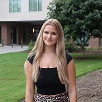 A smiling young woman with long blonde hair, wearing a black top and a patterned skirt, stands outdoors in front of a building and greenery.