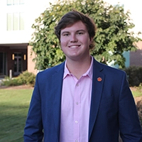 A young man in a blazer and collared shirt smiles confidently outdoors, with greenery in the background.