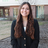 A smiling woman with long, wavy hair wearing a black blazer stands outdoors in front of a building.
