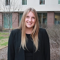 A smiling young woman in a black blazer standing outdoors in a professional setting.