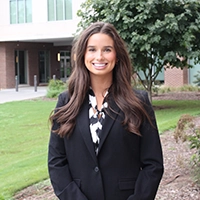 A smiling professional woman in business attire stands outdoors in front of a modern building.