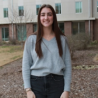 A young woman with long hair, wearing a gray sweater and black jeans, standing outside in front of a building.