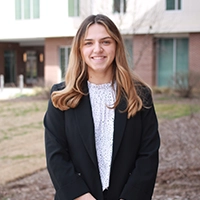 A smiling young woman in a black blazer stands outdoors in front of a modern building.