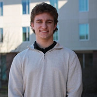 Young man smiling in a light sweater, standing outdoors in front of a modern building.