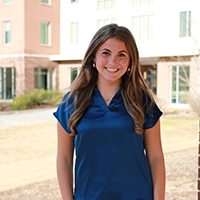 A young woman smiling while wearing a blue shirt, standing outdoors in front of modern buildings.