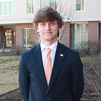 A professional portrait of a young man in a suit with a light orange tie, standing outdoors in front of a modern building.