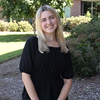 A smiling woman with shoulder-length blonde hair stands outdoors in a black dress, surrounded by greenery.