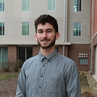 Photo of a young man with a beard standing outside a building, wearing a checked button-up shirt.