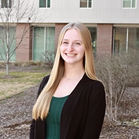 A young woman with long blond hair smiles outdoors, wearing a black cardigan over a green top, against a backdrop of a modern building.