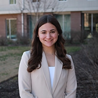 A young woman in a light-colored blazer smiles in an outdoor setting with a building in the background.