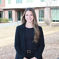 A young woman smiling in a professional outfit, standing outdoors in front of a modern building.
