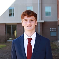 A young man in a suit with a red tie stands confidently in front of a modern building.