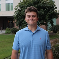 Smiling young man wearing a light blue polo shirt standing outside in front of a building.