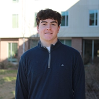 A young man standing outdoors in front of a building, smiling and wearing a navy blue sweater.