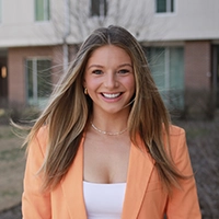 A smiling woman in a stylish orange blazer stands outdoors, showcasing a confident and approachable demeanor.