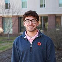A young man with curly hair and glasses wearing a navy blue sweatshirt smiles warmly against a backdrop of a modern building and greenery.