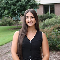 A smiling young woman with long brown hair, wearing a black sleeveless top, stands outdoors in a landscaped area.