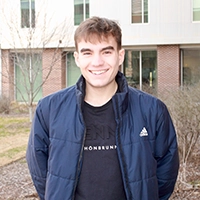 A young male smiling outdoors in a casual jacket, standing in front of a building with greenery.