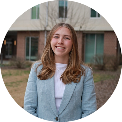 A young woman with long hair smiling, wearing a light blue blazer and standing outdoors in front of a building.