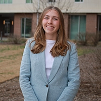 Portrait of a smiling young woman in a light blue blazer standing outside a building.