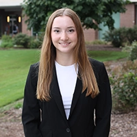 Professional headshot of a young woman wearing a black blazer in an outdoor setting.