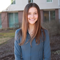 Headshot of a smiling young woman with long hair, wearing a blue jacket, set against a blurred outdoor background.