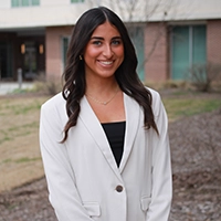 A young woman wearing a white blazer and black top stands outdoors, smiling in front of a building.