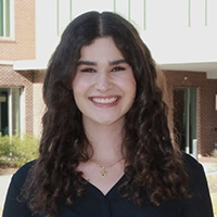 Portrait of a smiling young woman with long curly hair, dressed in a black top, standing outdoors.