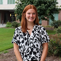 A smiling young woman with red hair wearing a black and white floral blouse stands outdoors in front of a building.