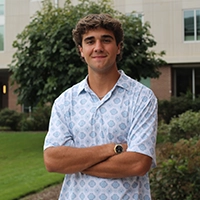 A young man standing confidently outdoors, wearing a patterned button-up shirt and smiling at the camera.