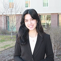 Professional portrait of a young woman in a black blazer, smiling outdoors with a building in the background.