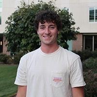 A smiling young man wearing a light-colored shirt stands outdoors in front of a building and greenery.